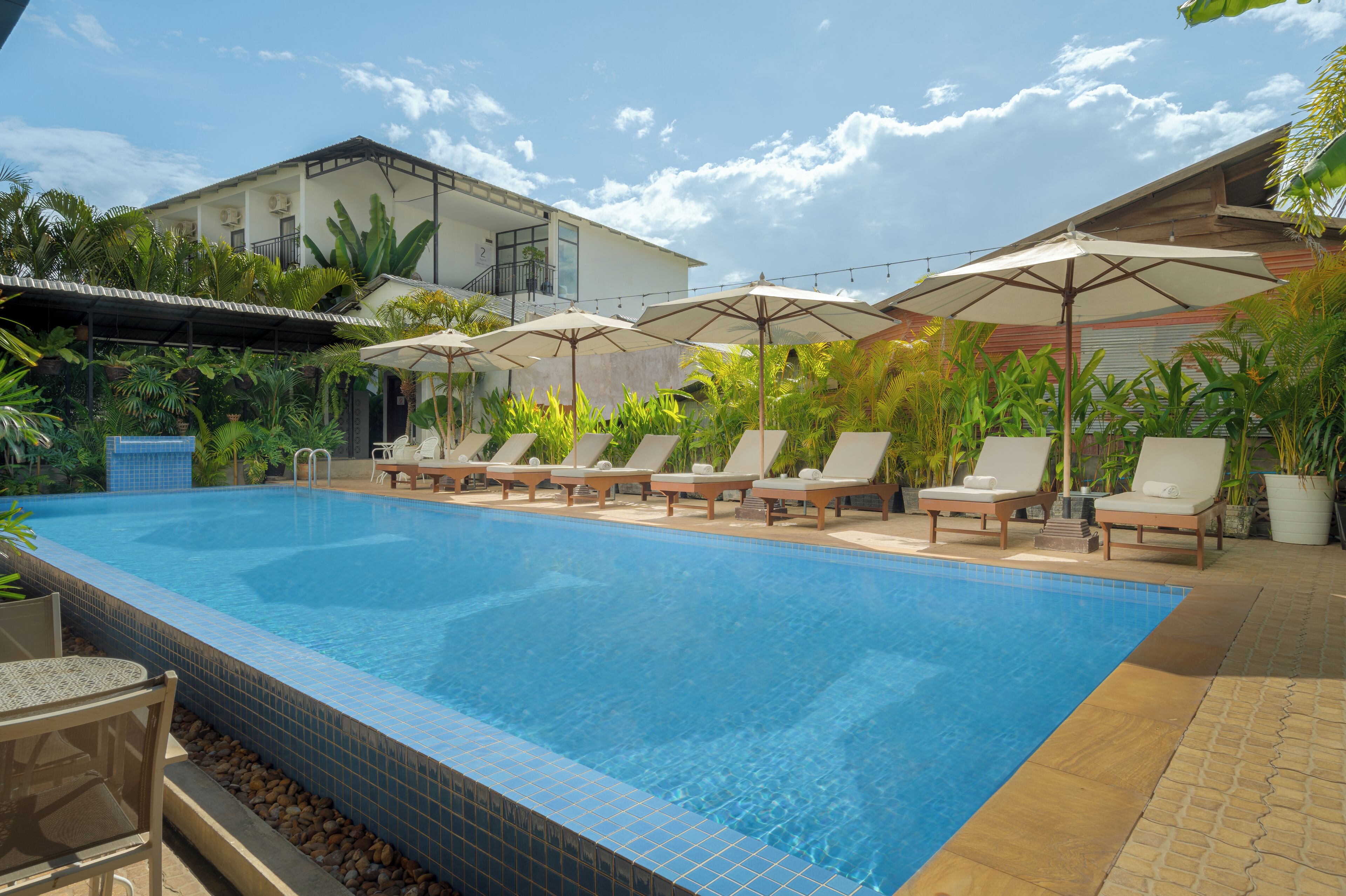 A swimming pool with lounge chairs and umbrellas, surrounded by greenery and a building with a balcony.