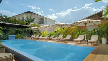 A swimming pool with lounge chairs and umbrellas, surrounded by greenery and a building with a balcony.