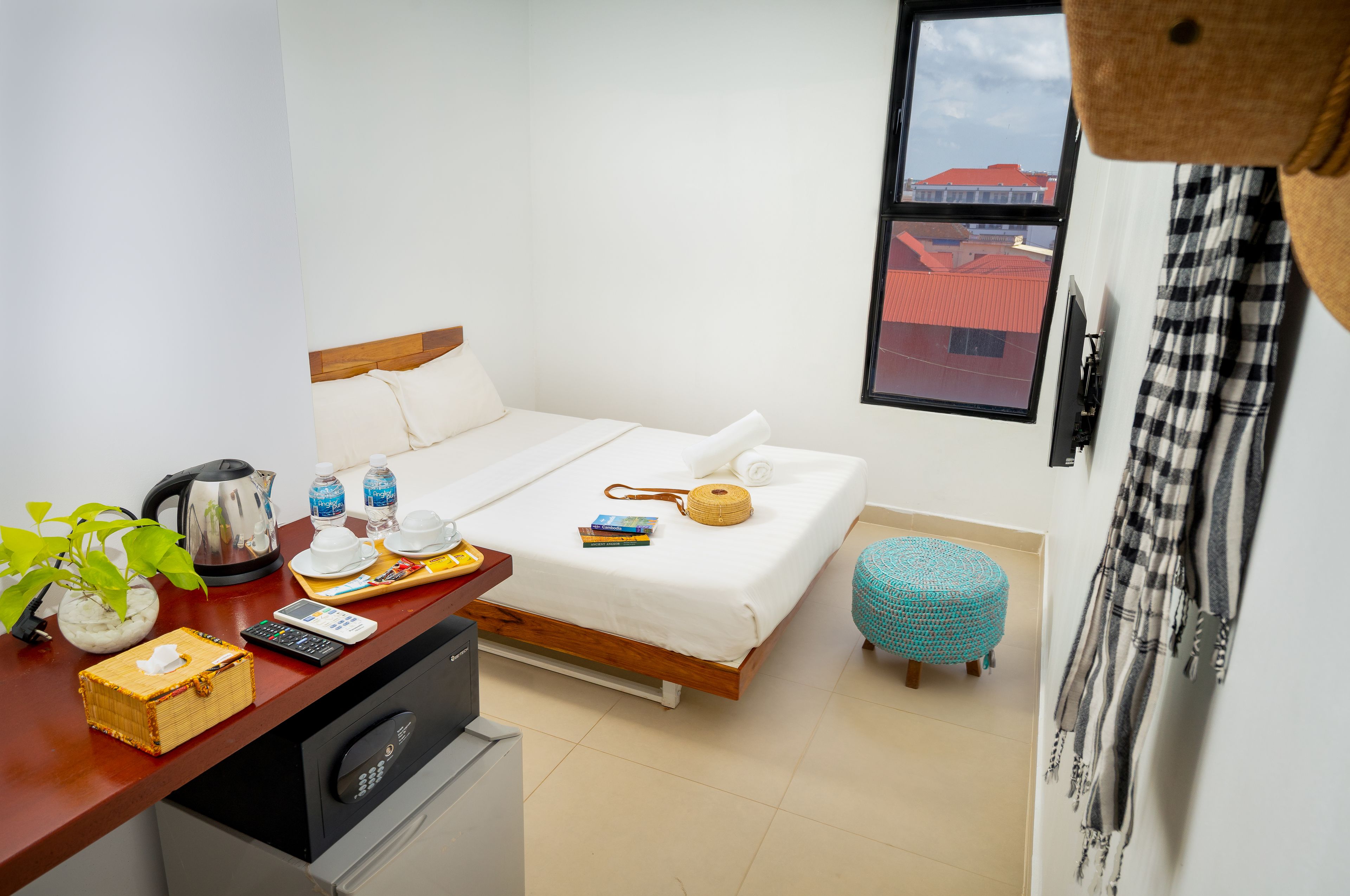 A neatly arranged hotel room with a bed, a small table with a kettle, a plant, and a window with a view of buildings.