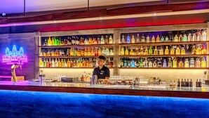 Una zona de bar con estantes llenos de botellas de licores, un letrero de neón y una barra con utensilios.