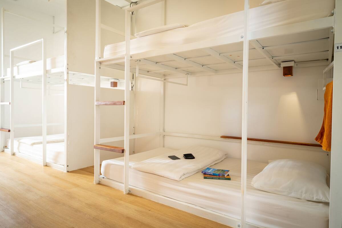 Habitación con literas de madera, suelo de madera, ropa de cama blanca y un libro en la litera inferior.