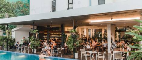 Uma área de piscina interna com assentos ao redor, um bar com pessoas e um edifício com vários andares e janelas.