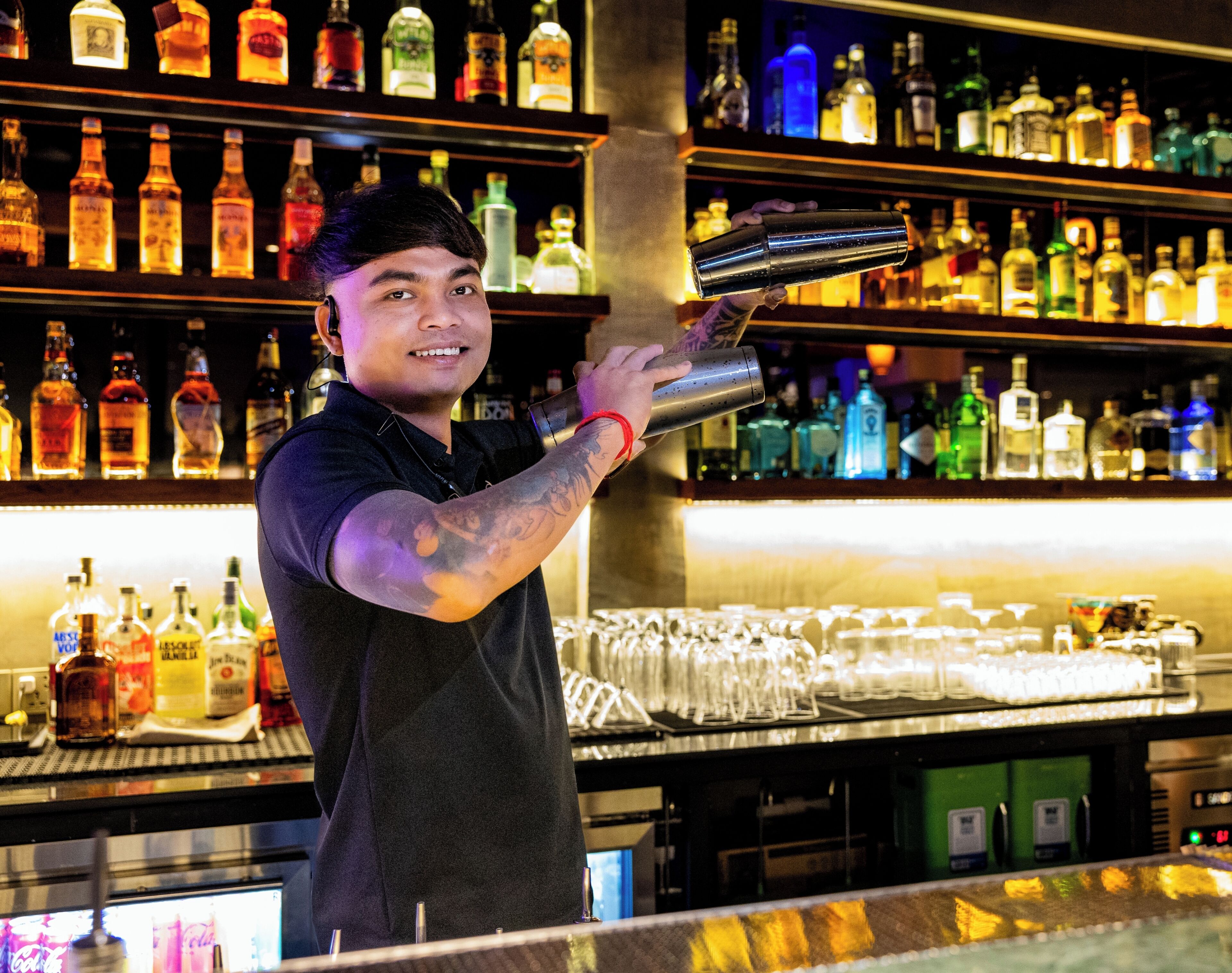A bartender holding a shaker at a bar with shelves of liquor bottles in the background.