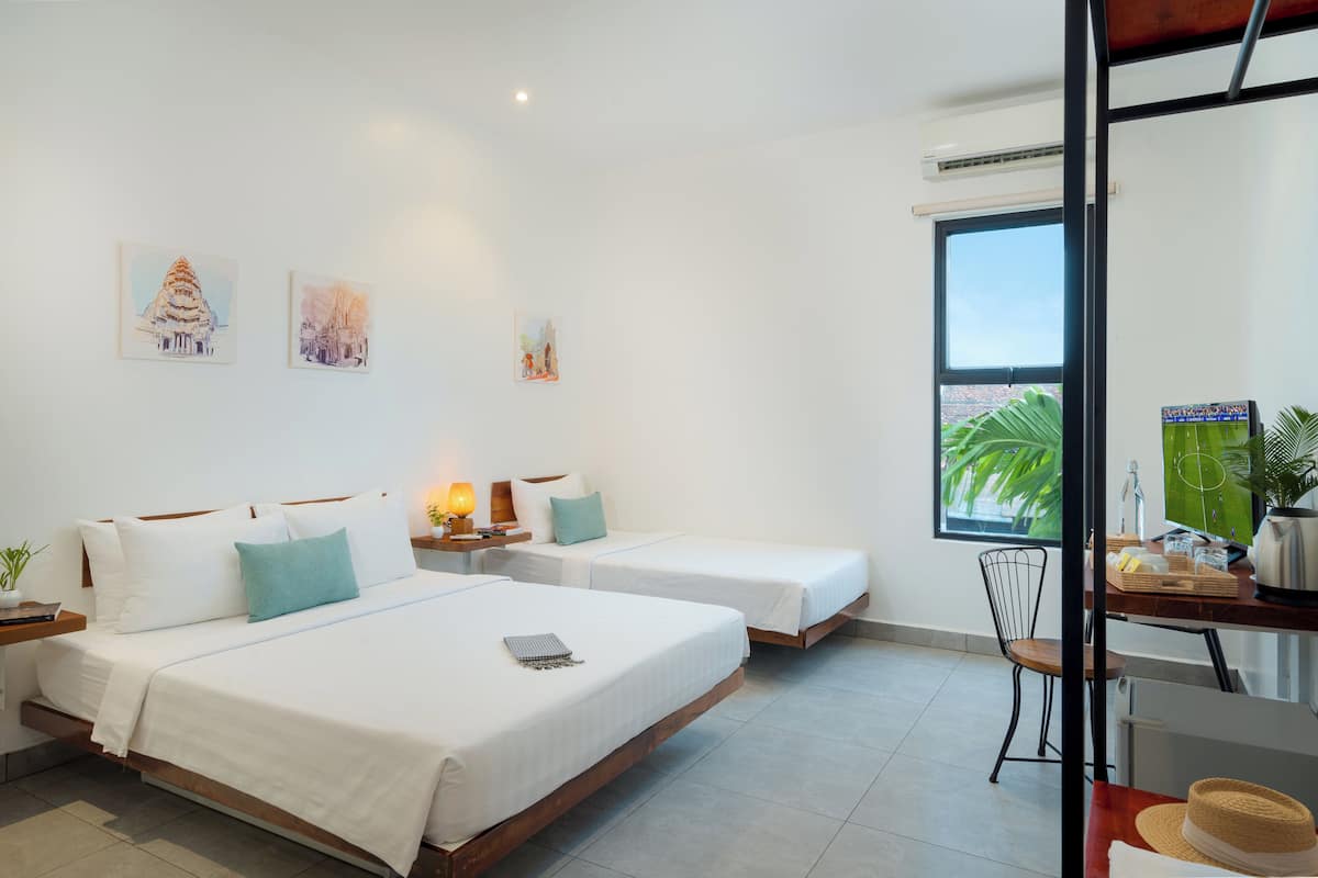 Habitación de hotel con dos camas, un escritorio con silla, una ventana con vista a la vegetación y una obra de arte en la pared.