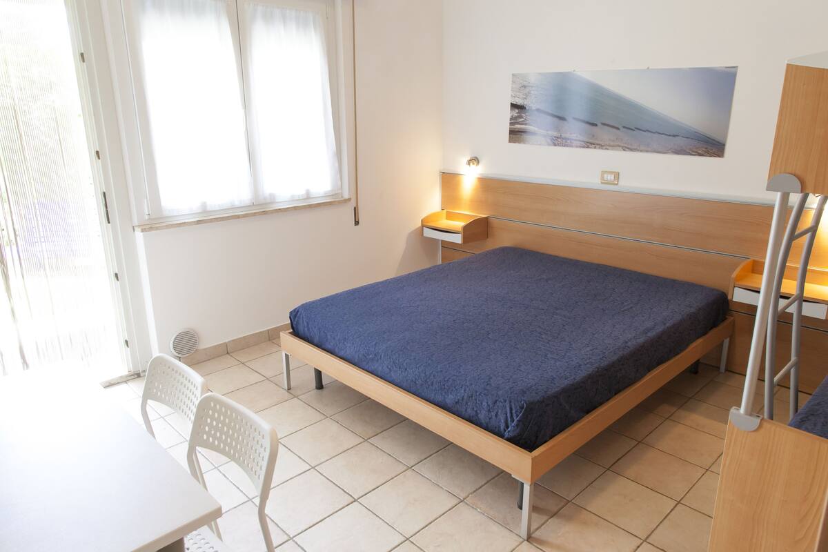 A bedroom with a bed, a desk, a chair, a nightstand, and a wall-mounted lamp.