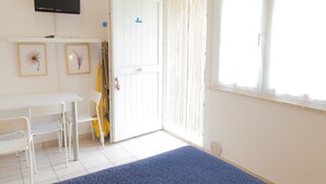 A room with a white desk, two chairs, a TV mounted on the wall, a door with a blue sign, and a window with sheer curtains. - Residence Casabianca (Fermo)
