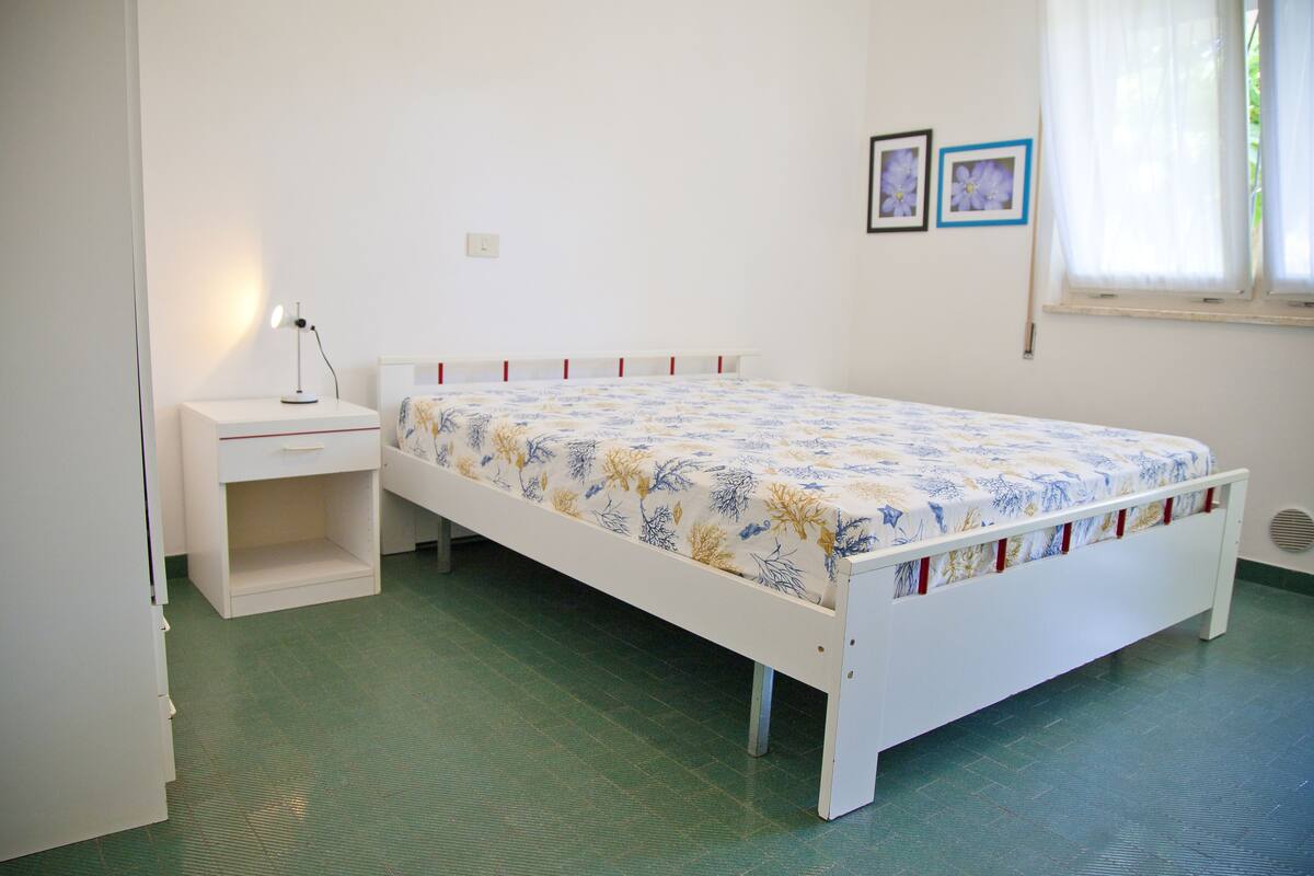 A single-bed room with a white bed, a nightstand, a small shelf, and a window with curtains.