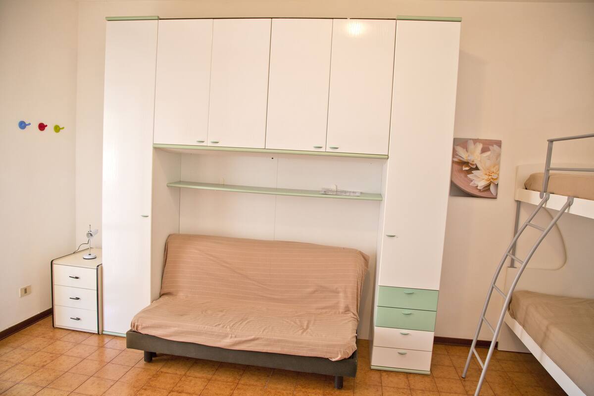 A bedroom with a bunk bed, a desk with drawers, a wardrobe with multiple shelves, and a small bedside table.