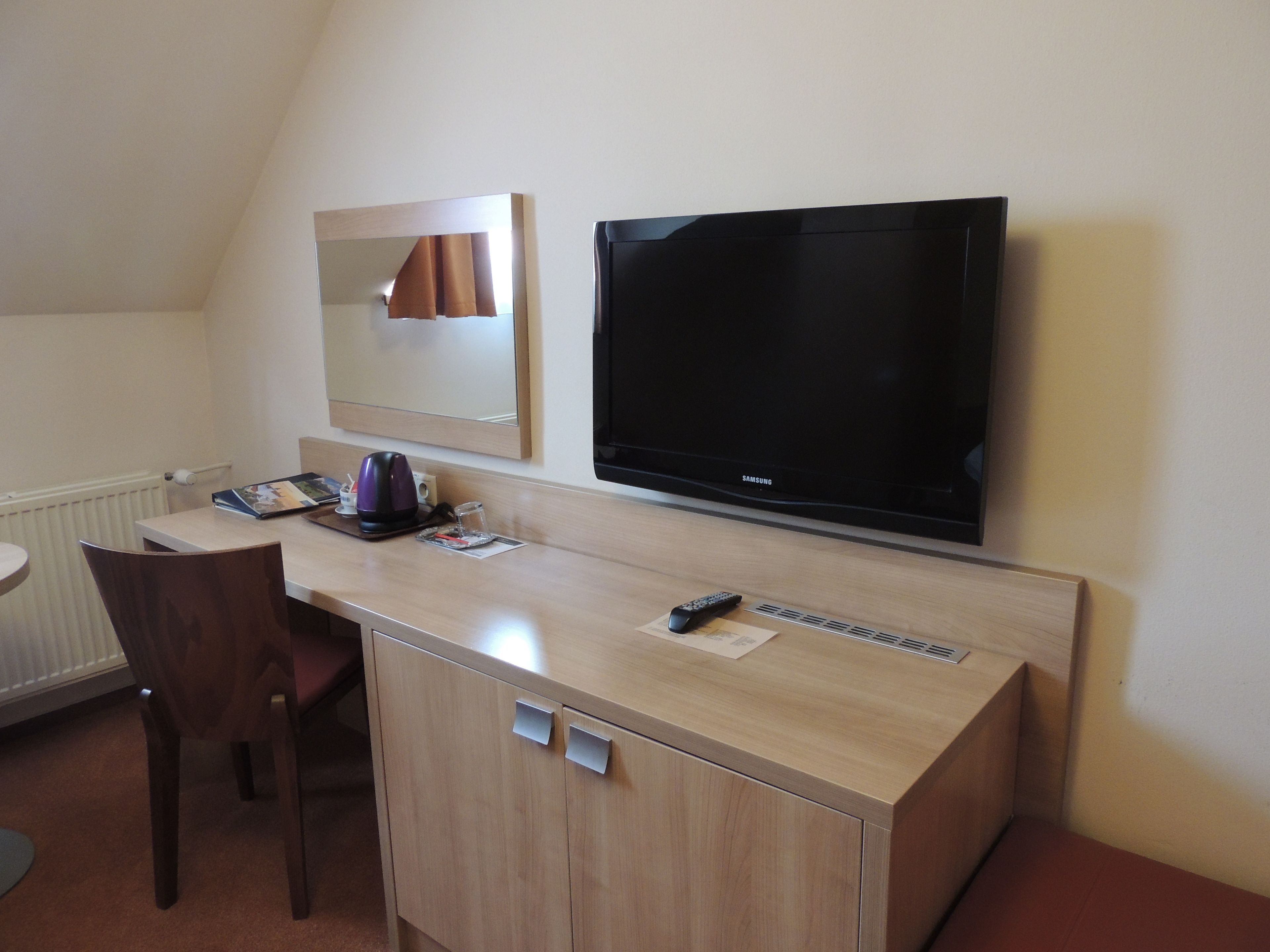 A hotel room with a wooden desk, a chair, a television, a mirror, and a radiator.