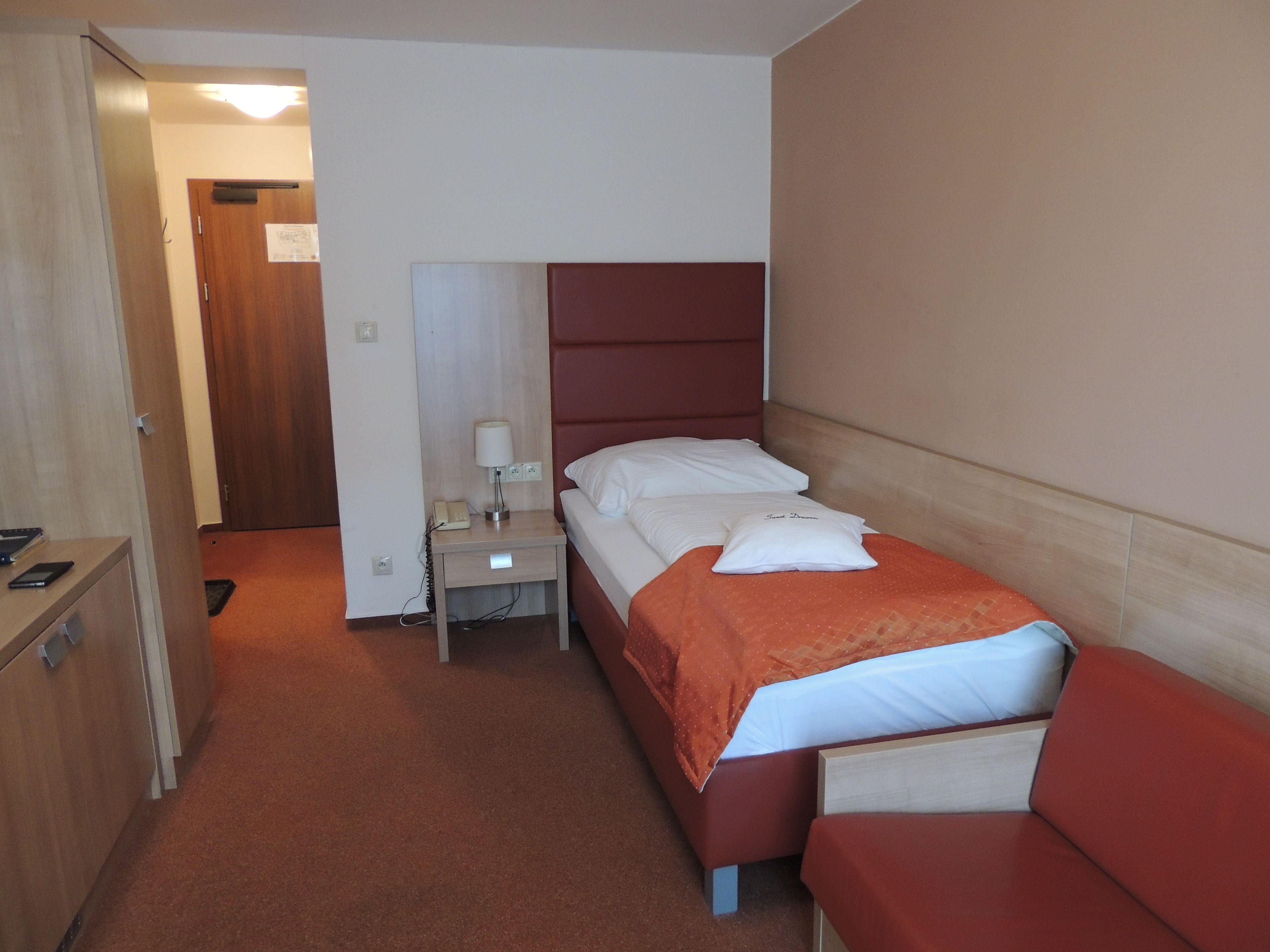 A hotel room with a single bed, a bedside table with a lamp, a small desk with a telephone, and a wardrobe.
