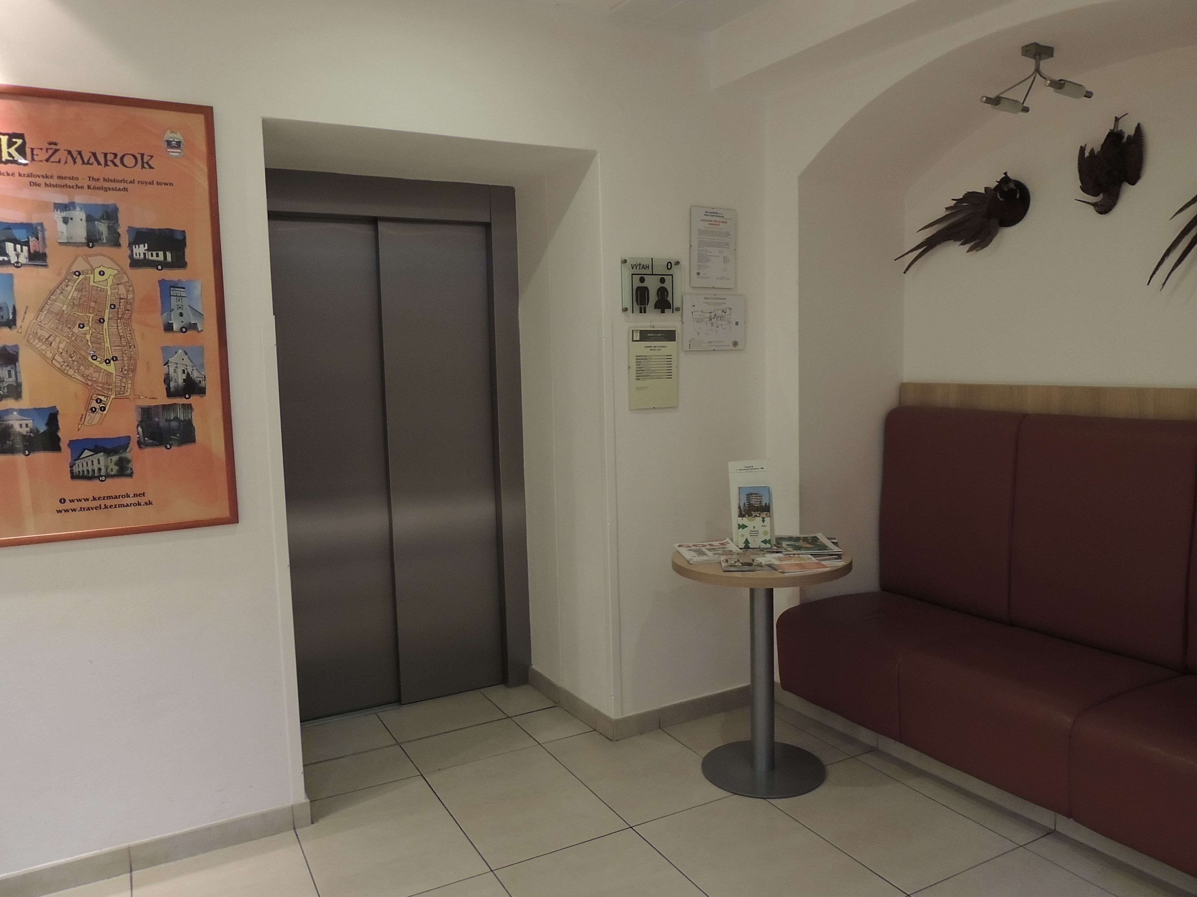 A hotel lobby with a red sofa, a small table with brochures, a poster of a historical town, and an elevator.