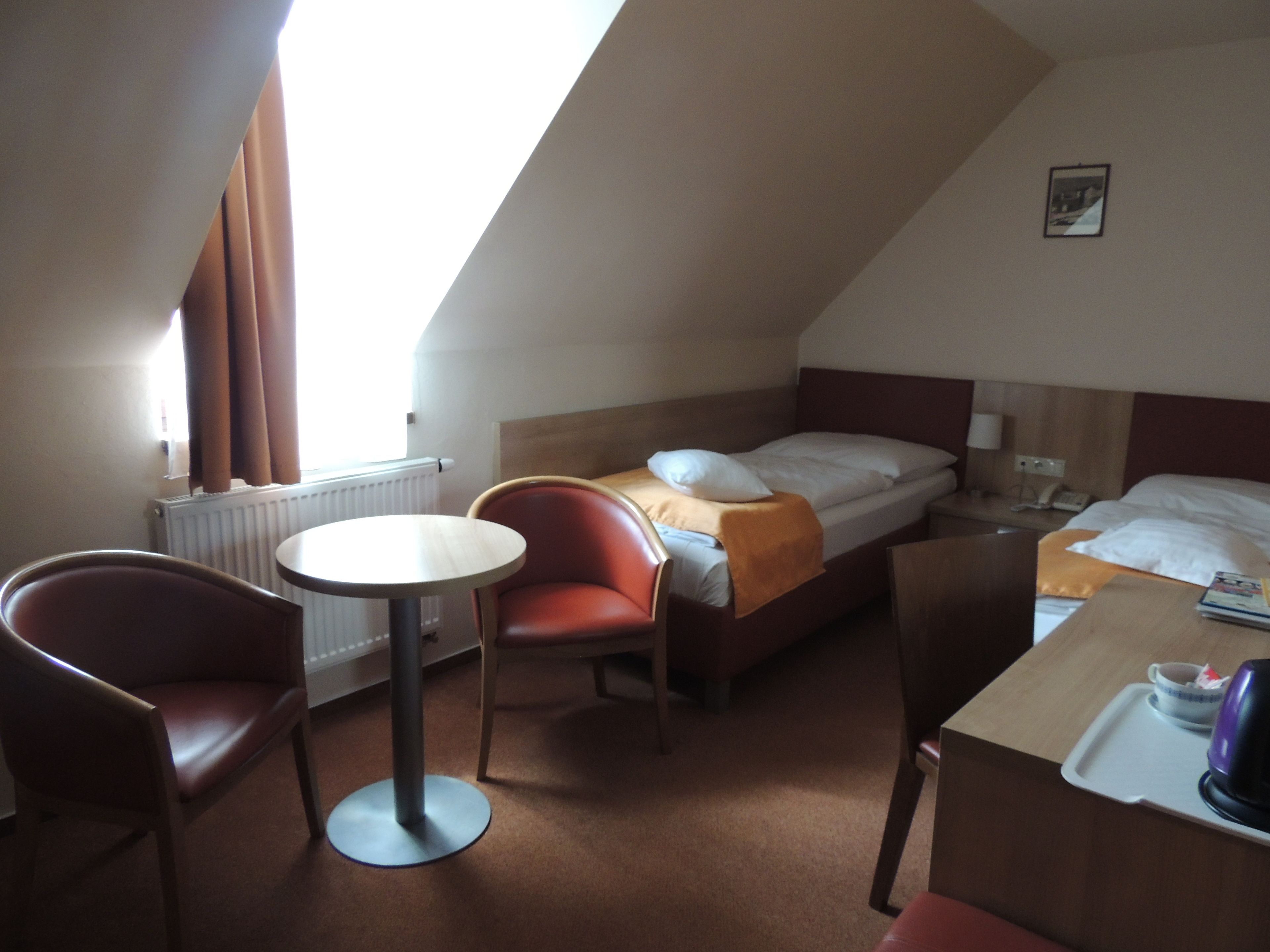 A hotel room with a bed, a desk, a chair, a table, and a radiator.