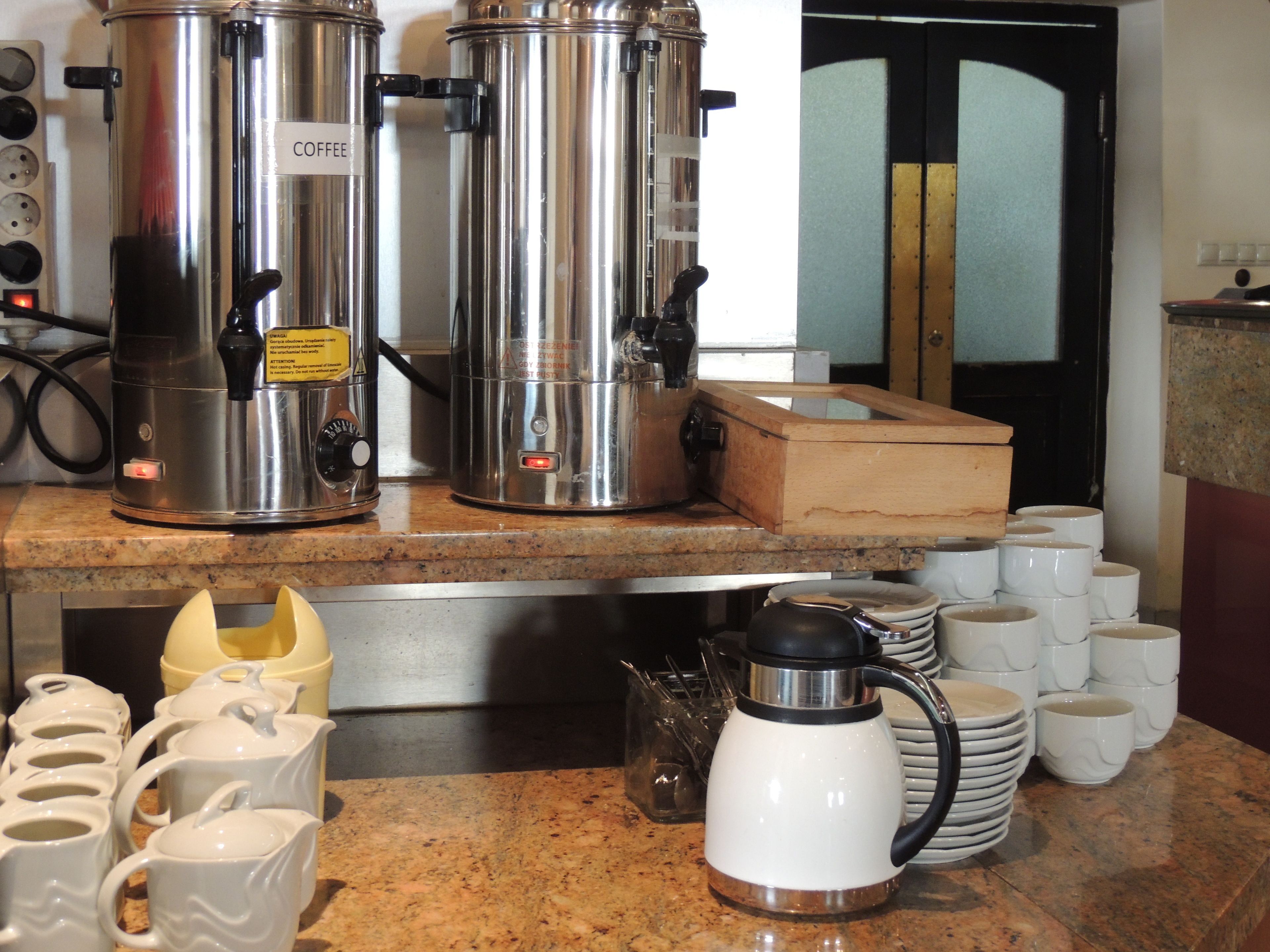 A kitchen area with stainless steel coffee makers, a coffee pot, and stacks of white mugs on the counter.