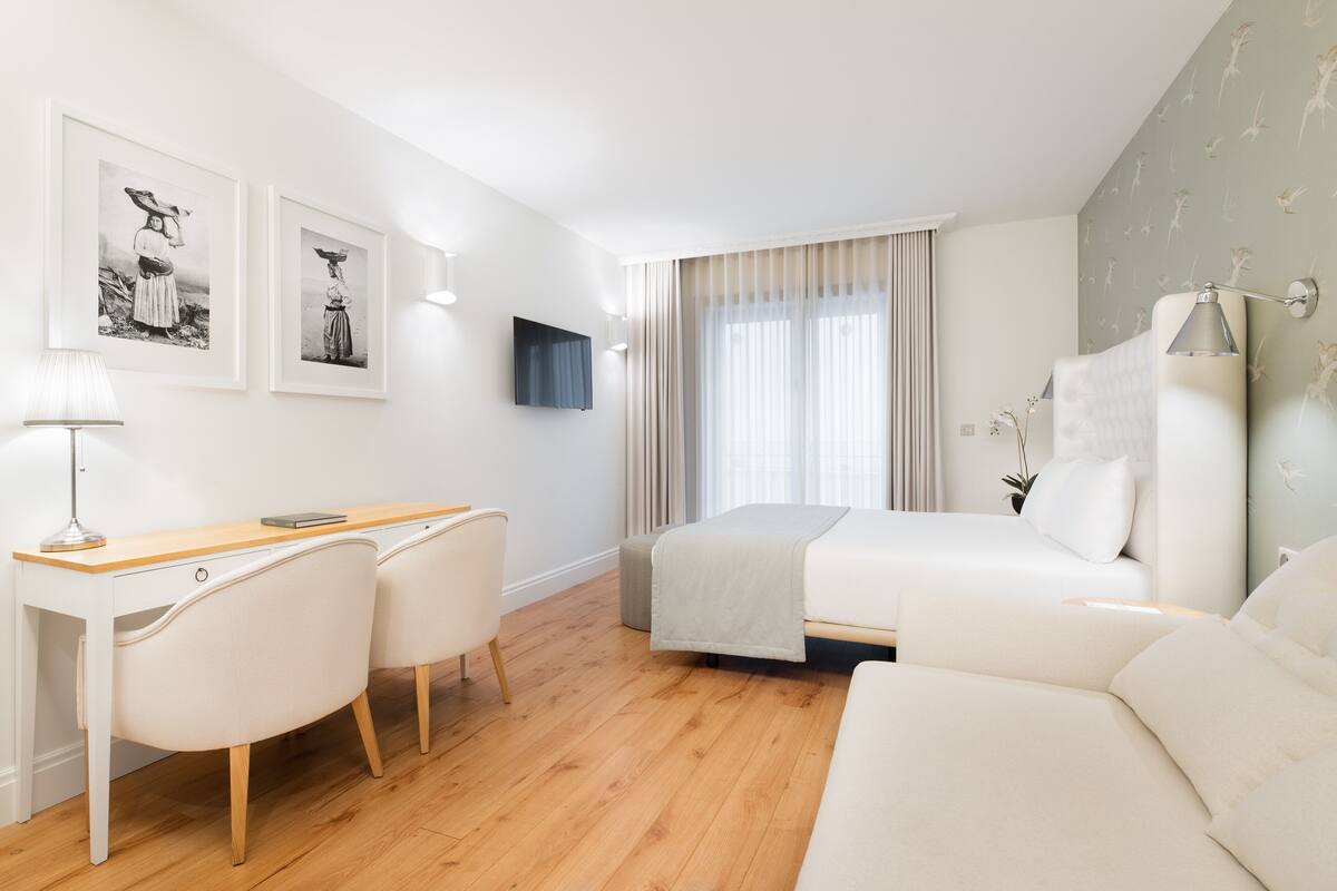 A modern hotel room with a wooden floor, a bed, a desk with a chair, and two framed pictures on the wall.