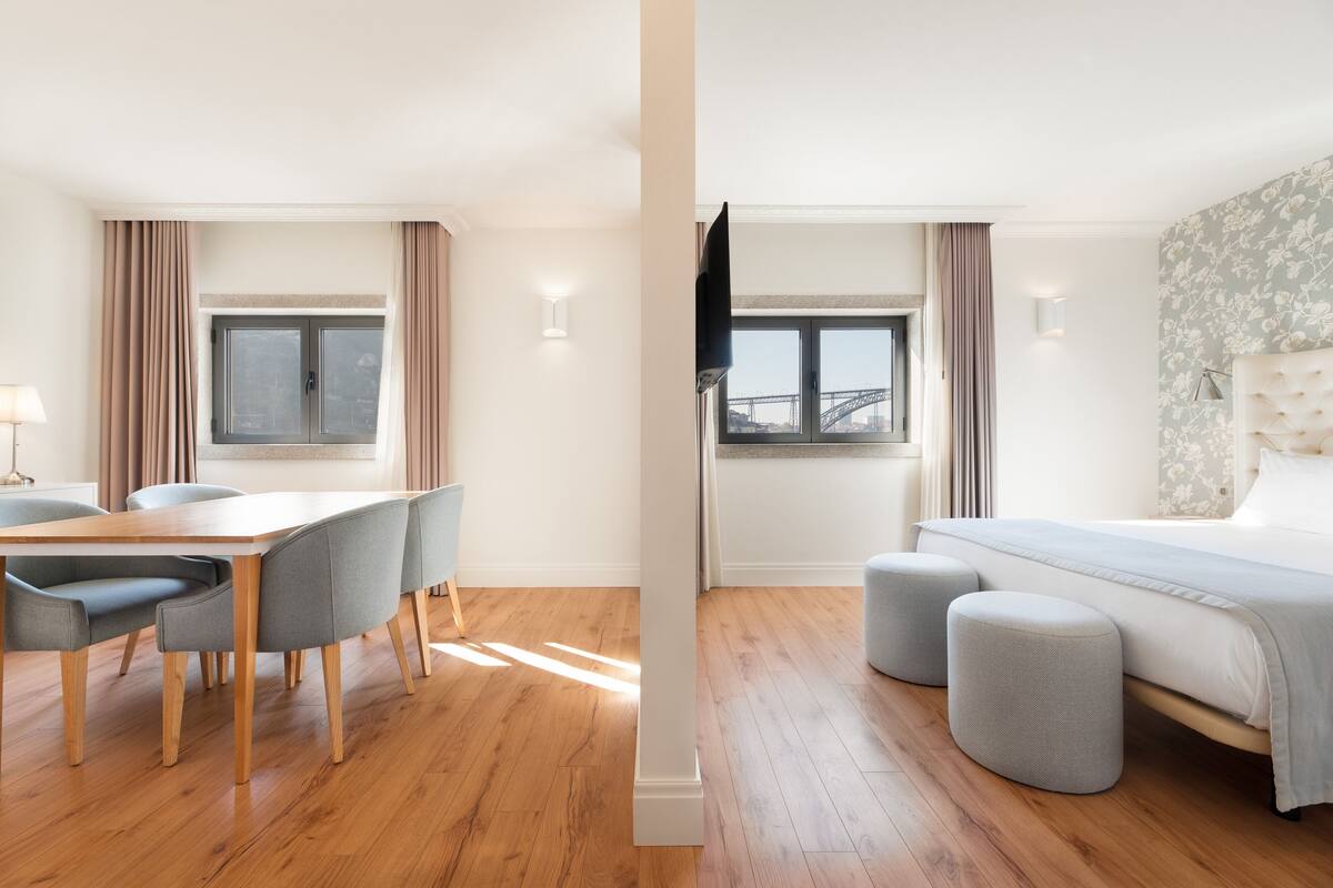 A modern hotel room with a wooden floor, a bed with grey ottomans, a dining area with a table and chairs, and a view of a bridge through the window.