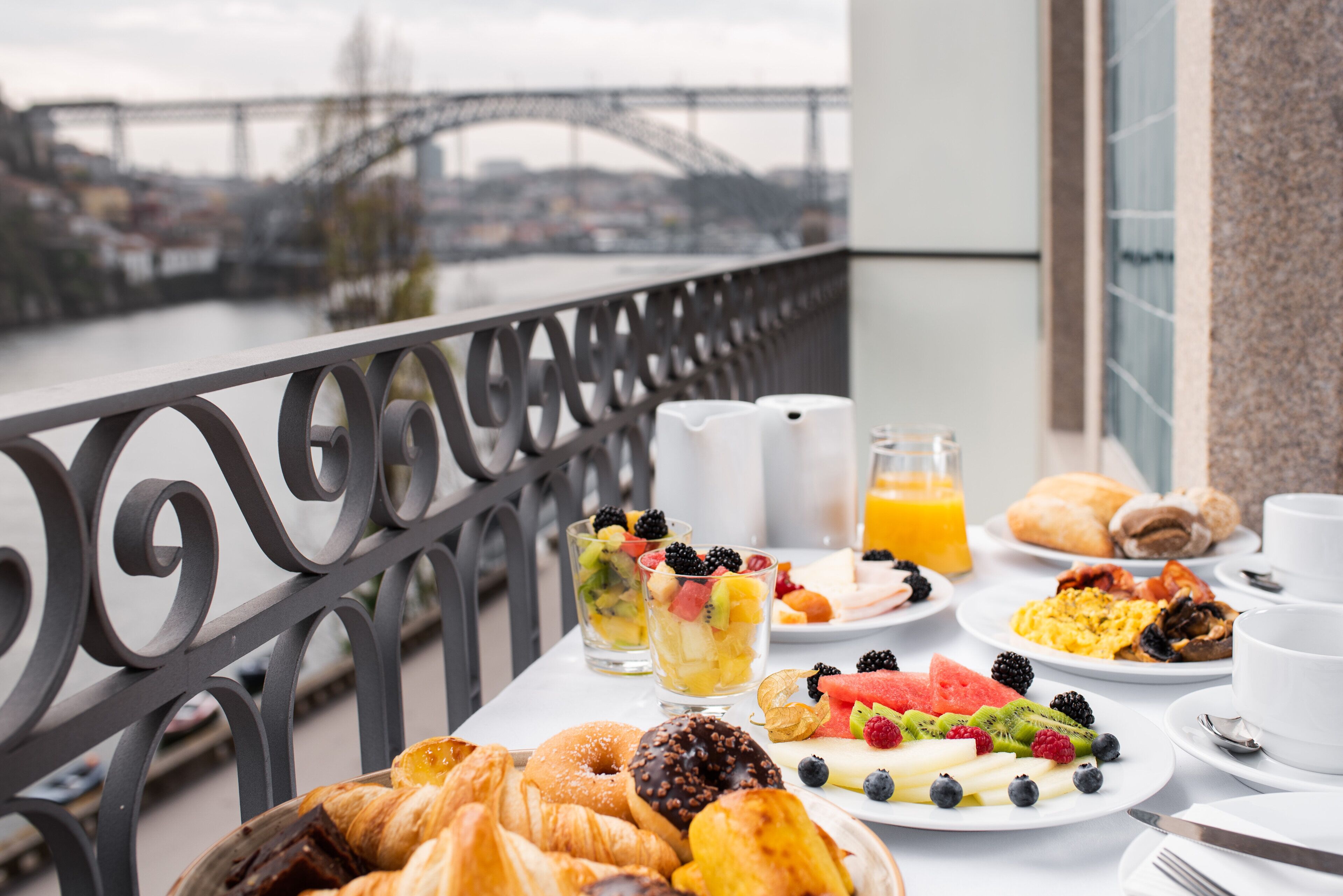 Ein Frühstückstisch mit Croissants, Obst und Getränken mit Blick auf einen Fluss und eine Brücke.