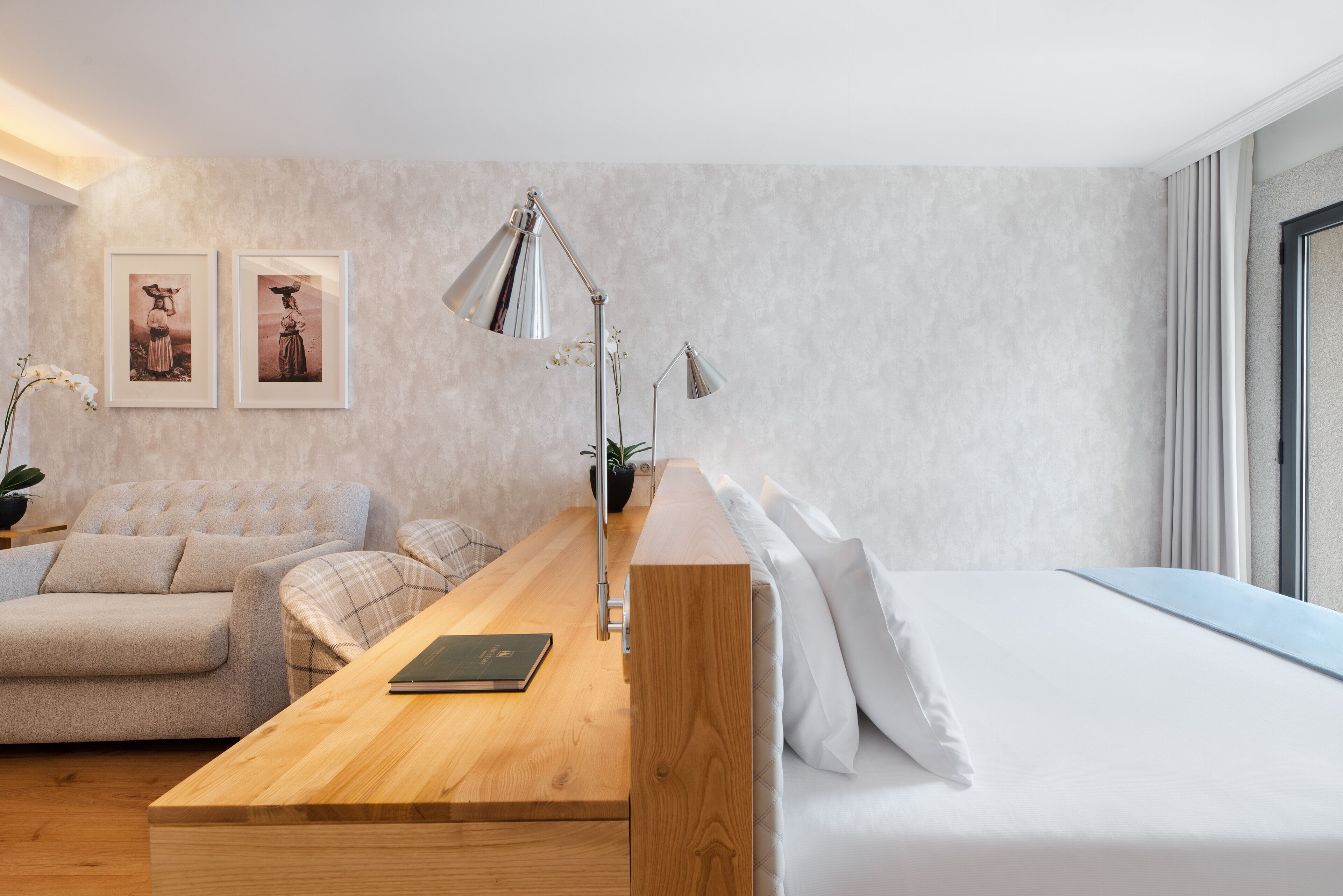 A modern hotel room with a wooden desk, a sofa, a bed, and two framed pictures on the wall.