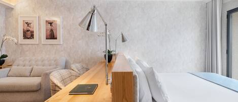 A modern hotel room with a wooden desk, a sofa, a bed, and two framed pictures on the wall.