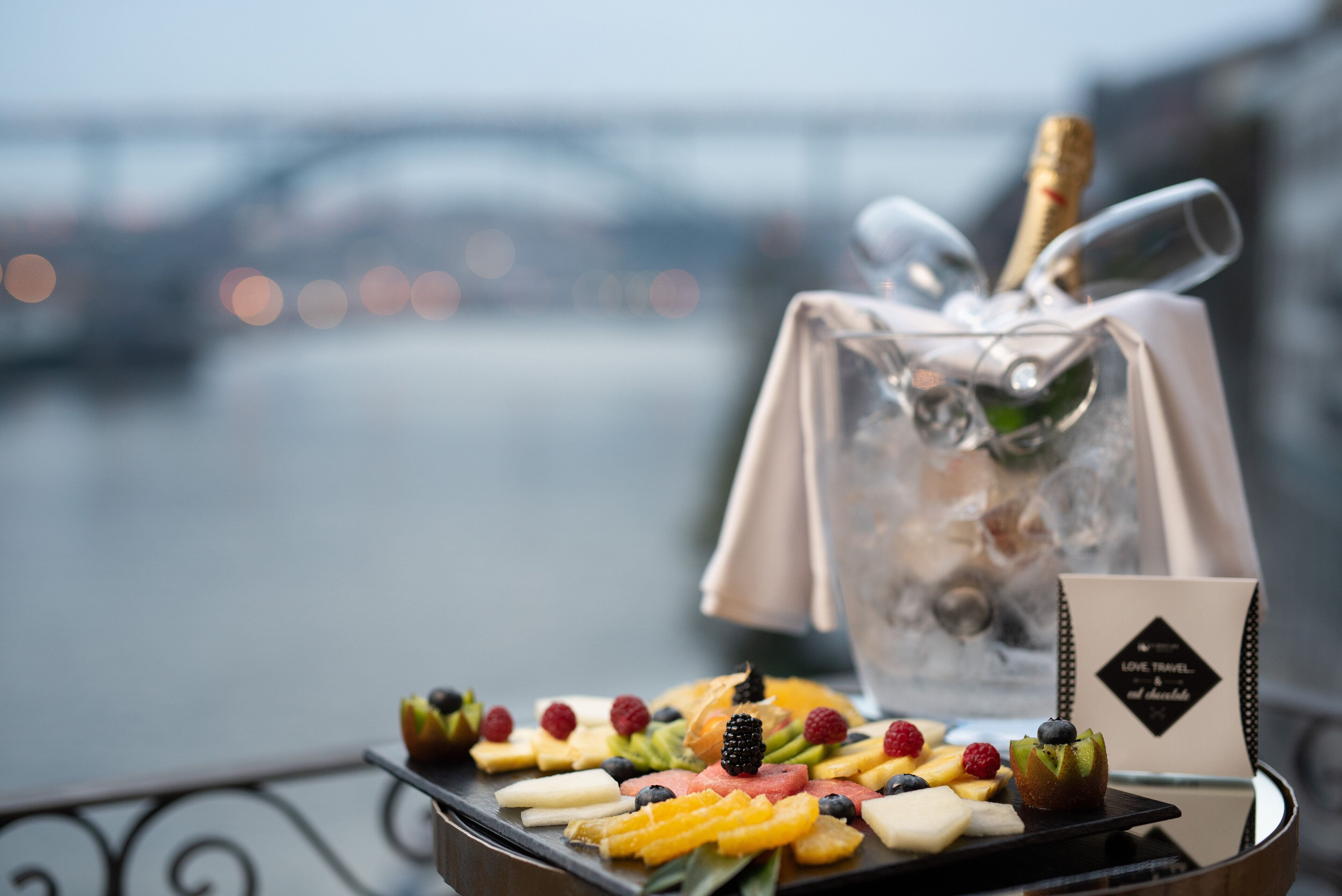 A fruit platter with a bottle of champagne and a menu on a balcony overlooking a river.
