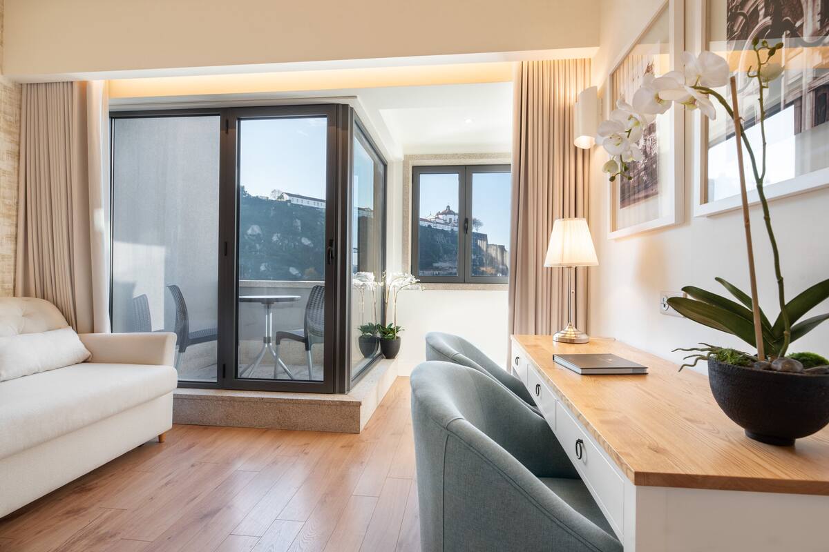 A modern hotel room with a sofa, a desk with a laptop, a potted orchid, and a view of a cityscape through large windows.