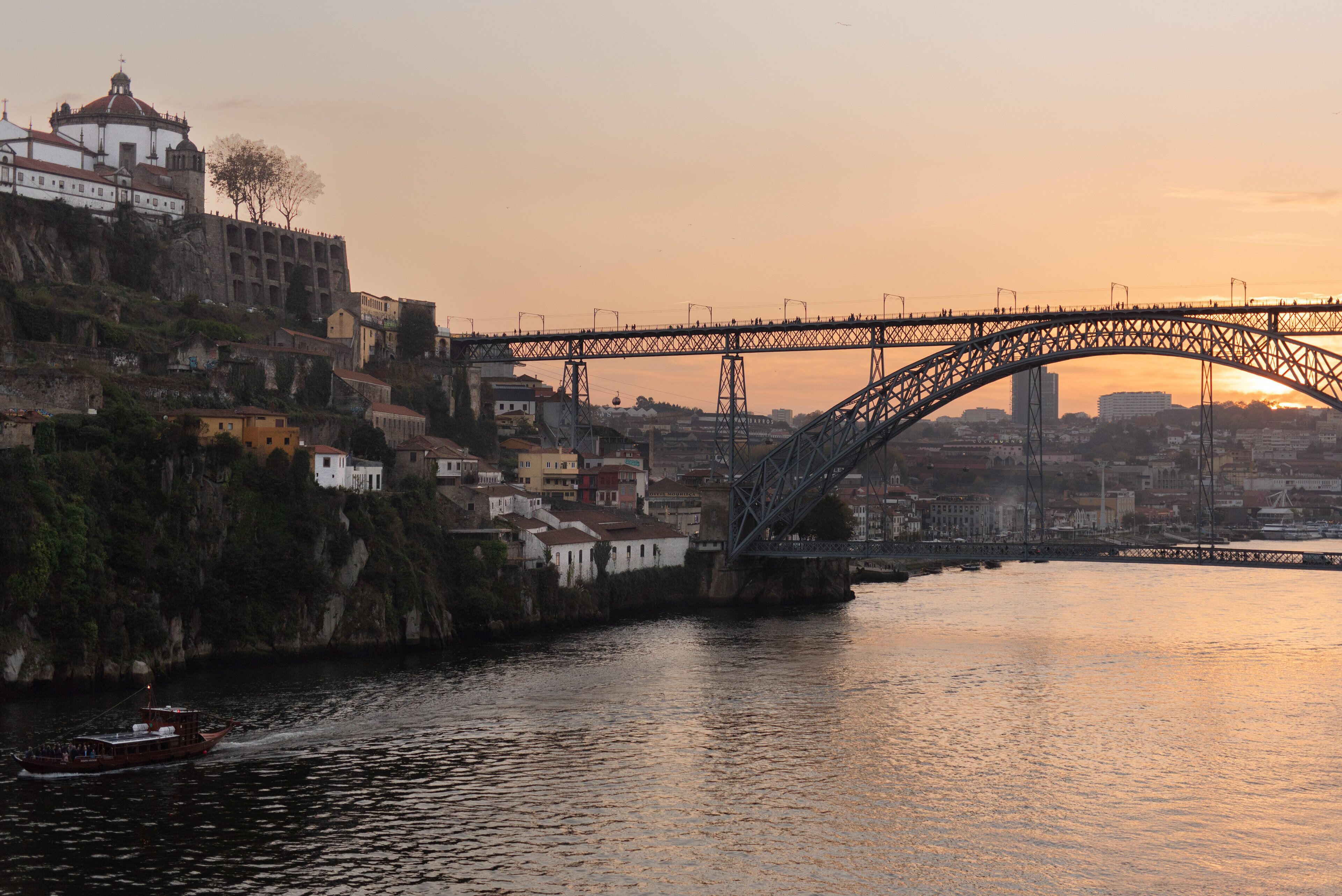 A bridge over a river with buildings on the left and a sunset in the background.