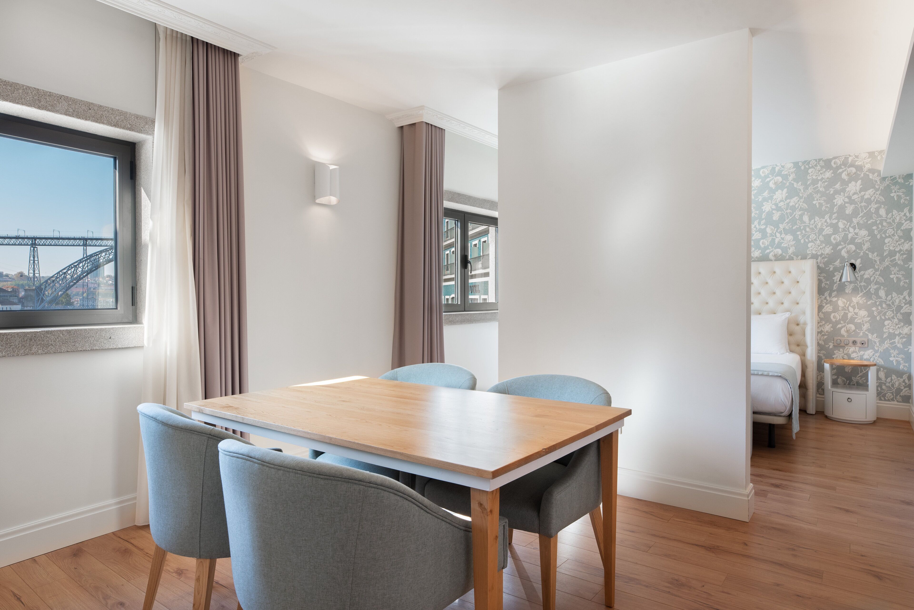 A modern room with a wooden dining table, four chairs, a bed with a white headboard, a nightstand, and a view of a bridge through a window.