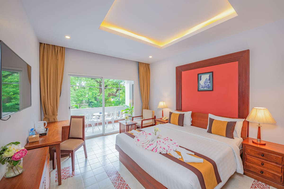 A hotel room with a large bed, a desk with a chair, a small table with chairs, a window with curtains, and a balcony with a table and chairs.