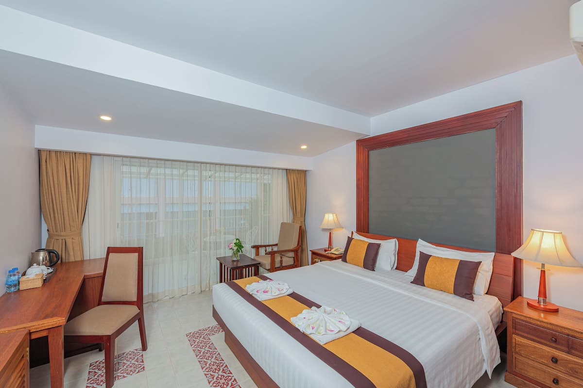 A hotel room with a large bed, a desk with a chair, a small table with a lamp, and a window with curtains.