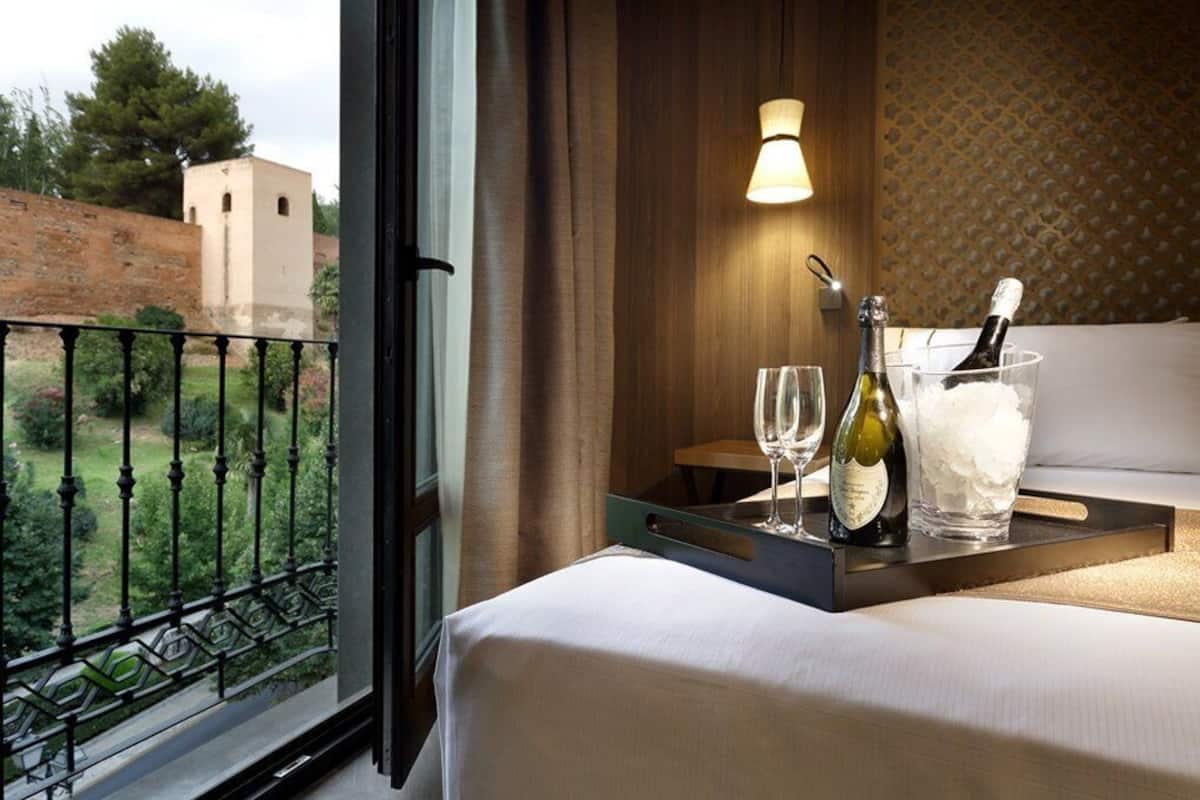 Ein Hotelzimmer mit Balkon, einem Bett, einem Nachttisch mit einer Weinflasche, zwei Weingläsern und einem Kerzenhalter.