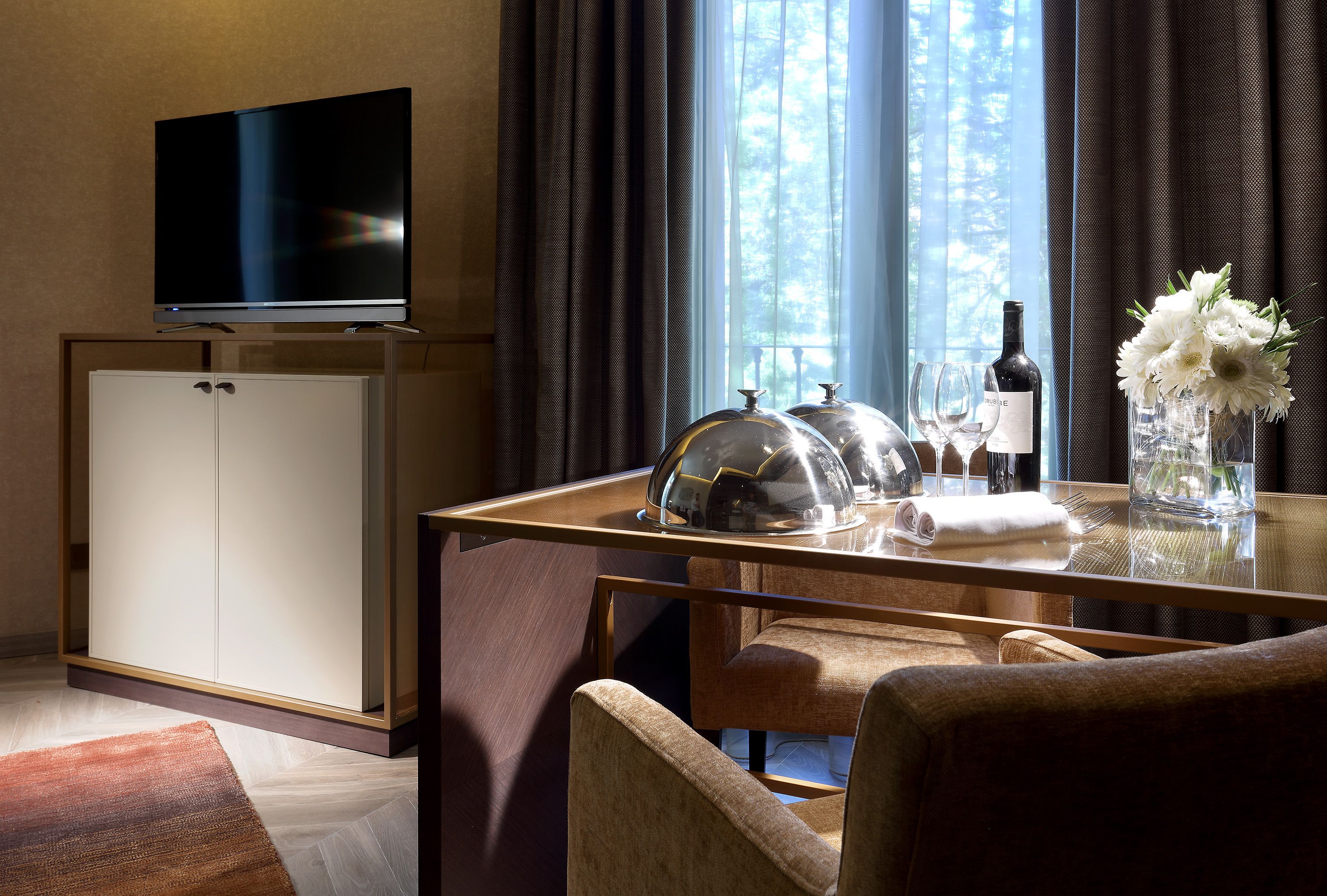 Ein modernes Hotelzimmer mit Fernseher, Minibar, einem Esstisch mit Glasplatte, einer Couch und Blick auf Bäume durch das Fenster.