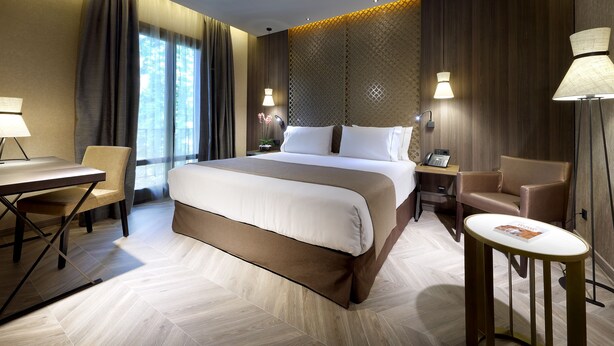 A modern hotel room with a large bed, a desk, a chair, and a small table.