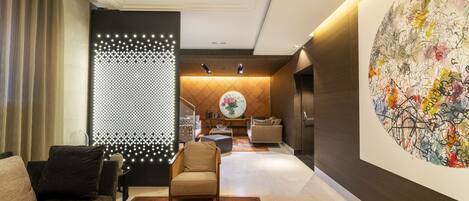 Un moderno vestíbulo de hotel con un sofá, un sillón y una mesa de centro. Se observa una pared decorativa con luces y un cuadro abstracto de gran tamaño.