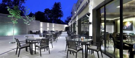 An outdoor dining area with tables and chairs, a glass wall, and a building in the background.