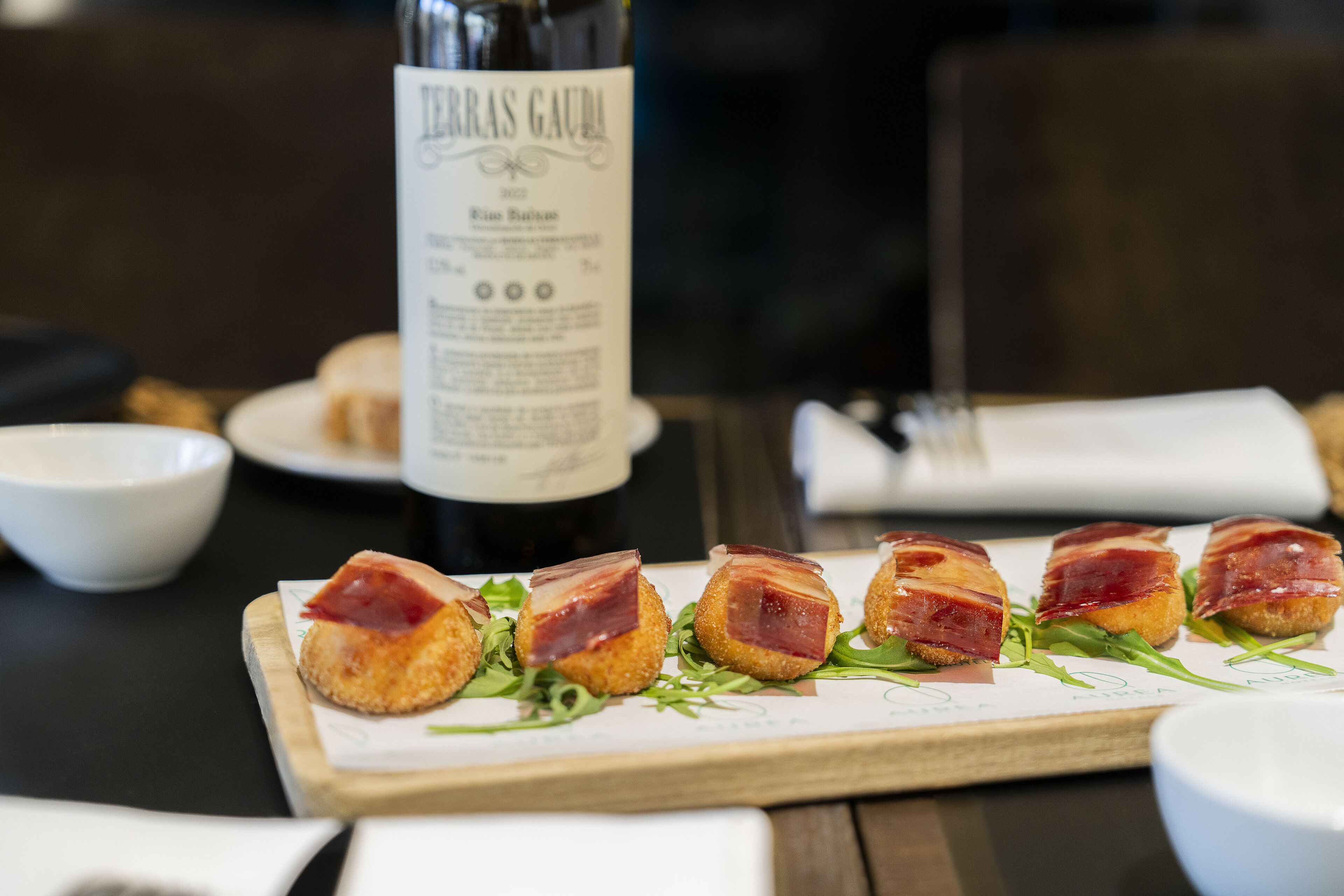 A table with a dish of bacon-wrapped items, a bottle of wine, and a white bowl.