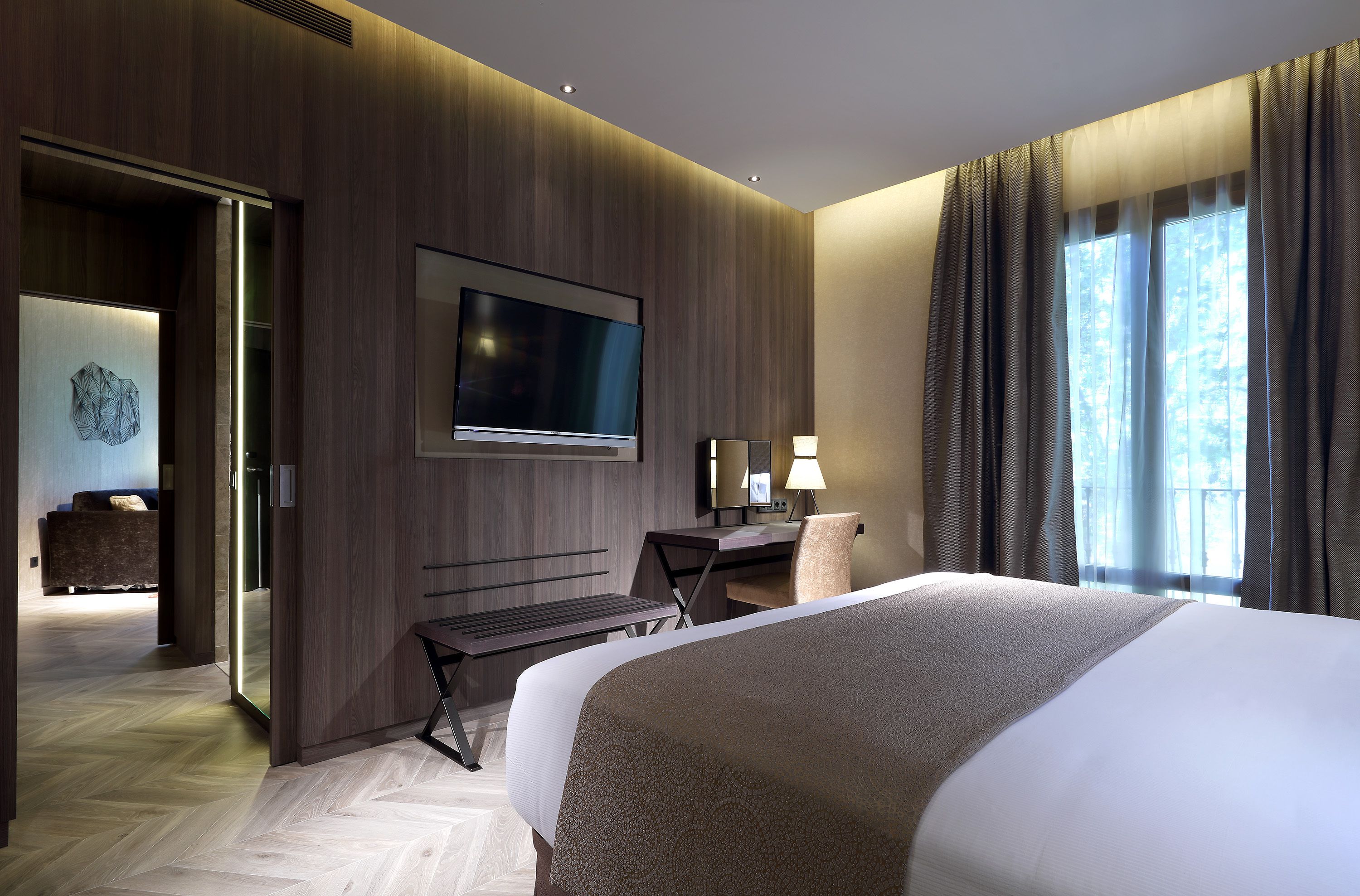 A modern hotel room with a large bed, a desk, a flat-screen TV, and a view of the outdoors through the window.