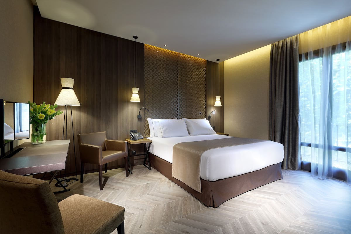 Ein Hotelzimmer mit einem großen Bett, einem Schreibtisch mit Stuhl, einer Lampe und Blick ins Freie durch ein Fenster.