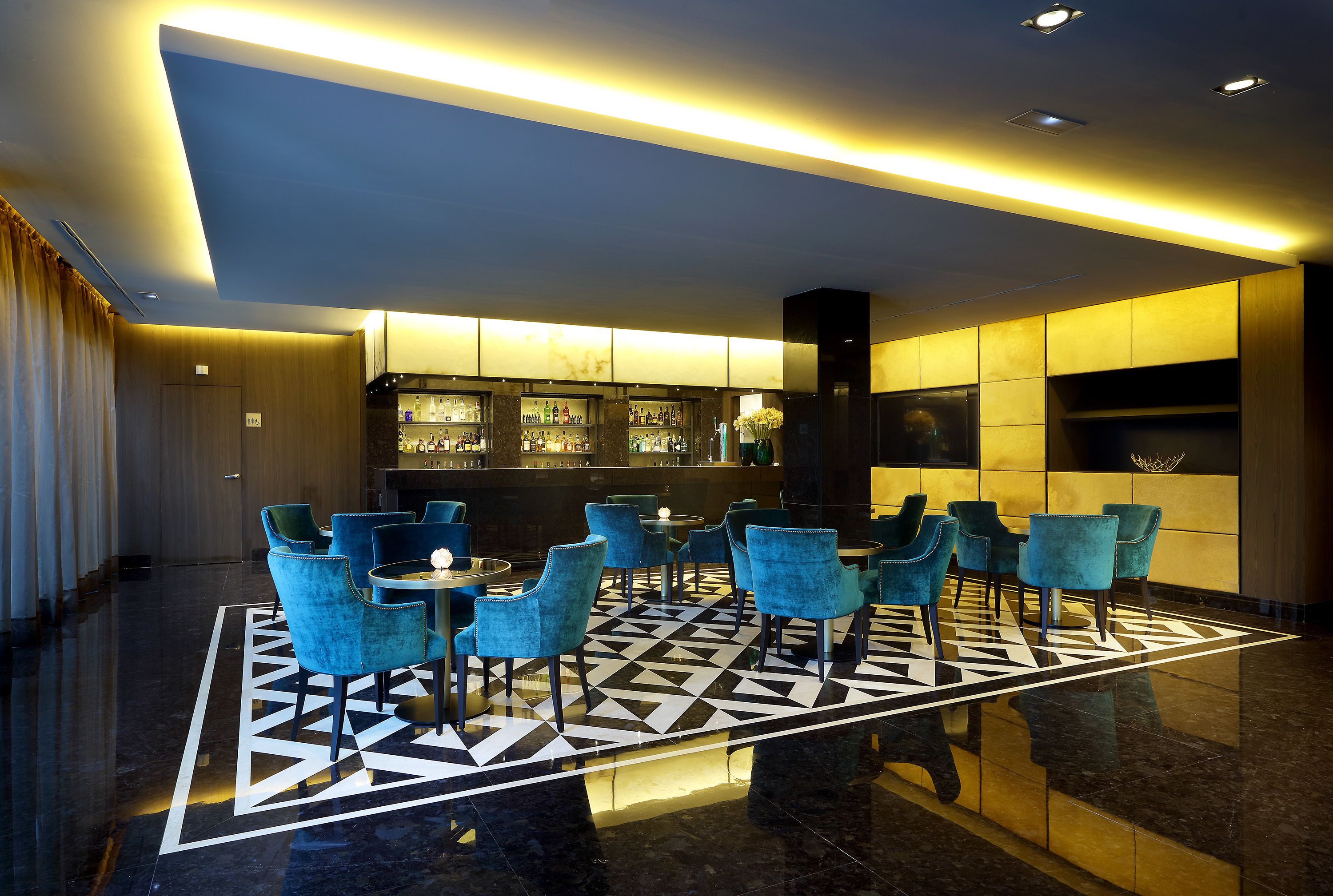 A lobby with a geometric patterned floor, blue chairs, a bar area, and a modern ceiling design.