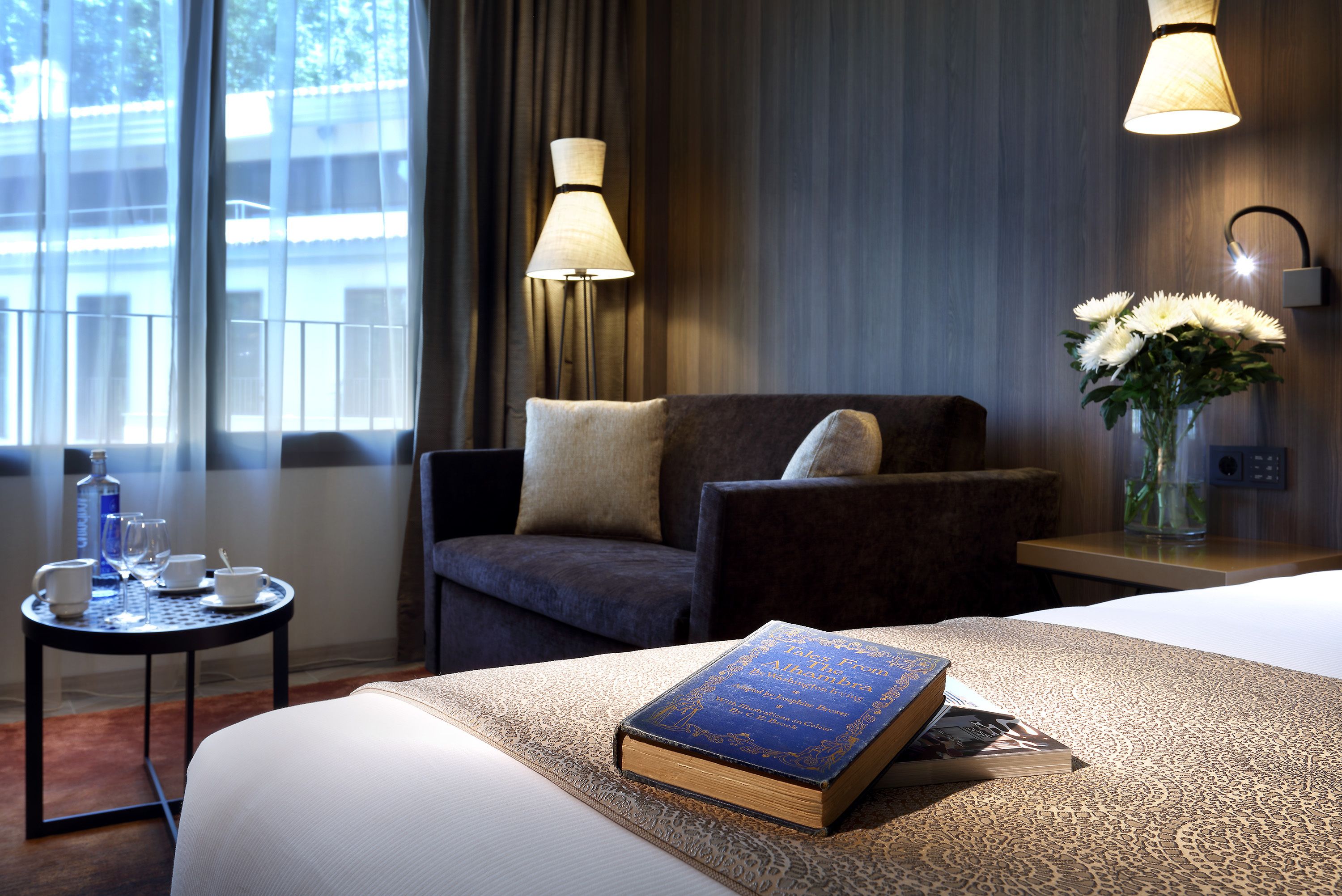 A hotel room with a bed, a chair, a small table with a book, a vase with flowers, and a lamp.