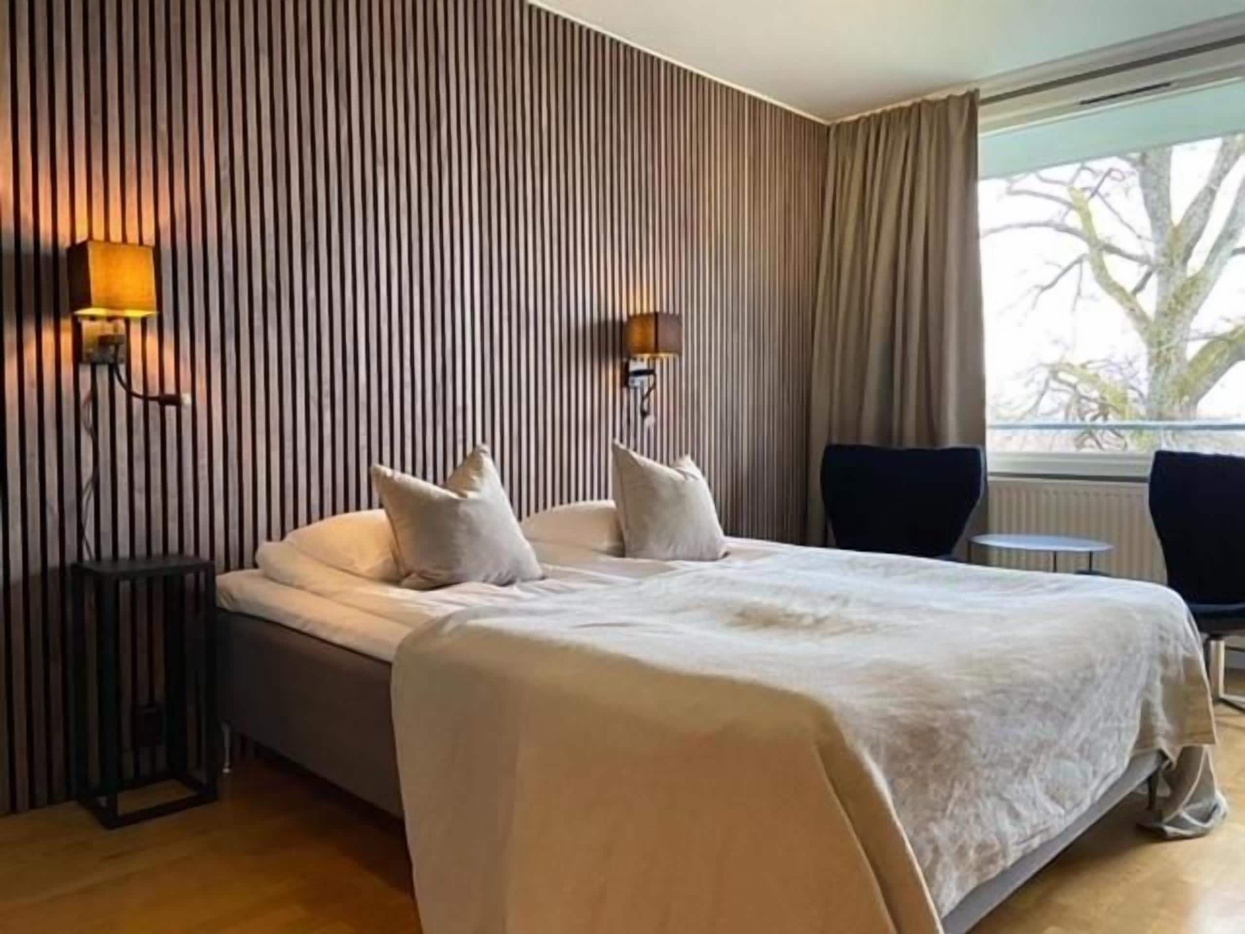 Een hotelkamer met een groot bed, twee bedlampen, een raam met gordijnen en uitzicht op de omgeving.