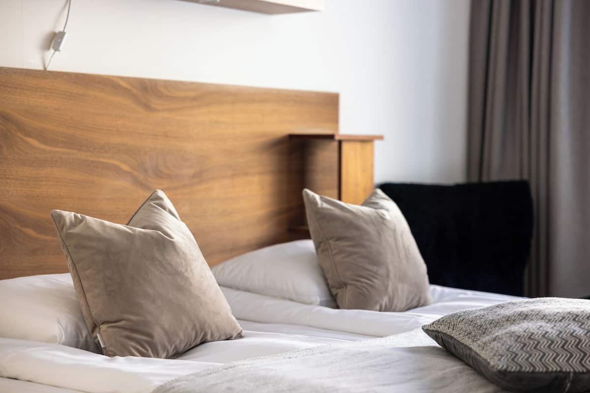 Una cama bien hecha con sábanas blancas y almohadas color beige, un cabecero de madera y una almohada oscura a la derecha.