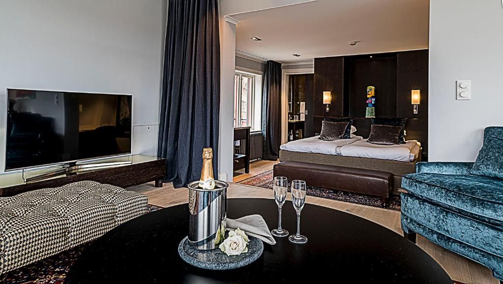 A hotel room with a bed, a TV, a dining table with a bottle and glasses, and a blue armchair.