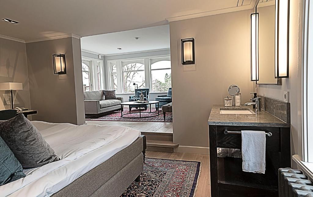 A hotel room with a bed, a sitting area with a sofa and chairs, a bathroom with a mirror and sink, and large windows.