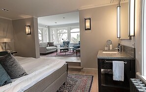 Une chambre d’hôtel comprenant un lit, un coin salon avec un canapé et des fauteuils, une salle de bain avec un miroir et un lavabo, et de grandes fenêtres.