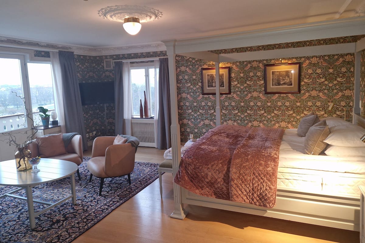Una habitación de hotel con una cama, dos sillones, una mesita y un ventanal grande.