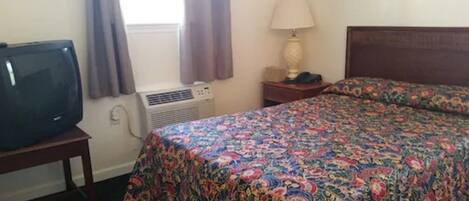 A hotel room with a bed, a TV, an air conditioner, a lamp, and a window with curtains.