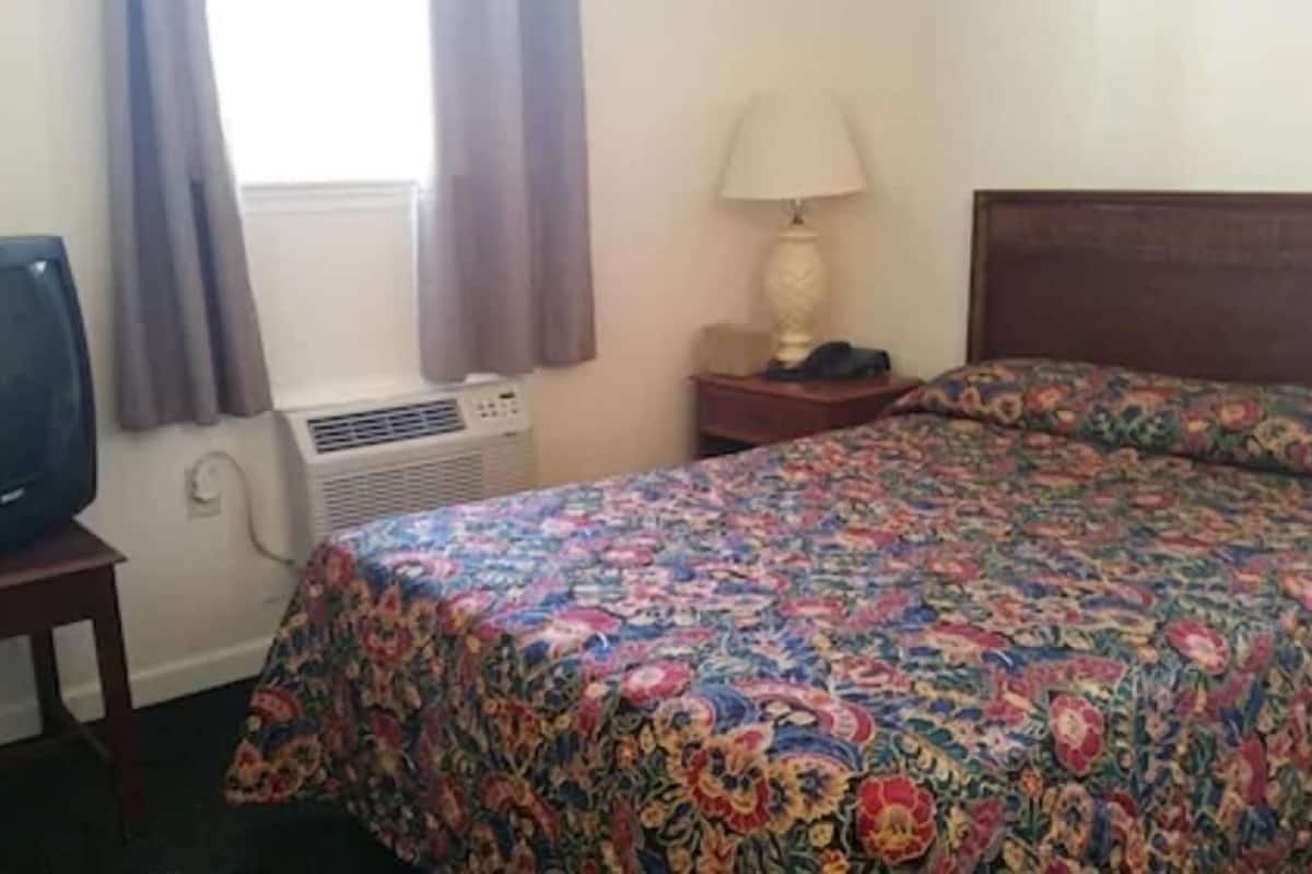 A hotel room with a bed, a television, an air conditioning unit, a lamp, and a window with curtains.