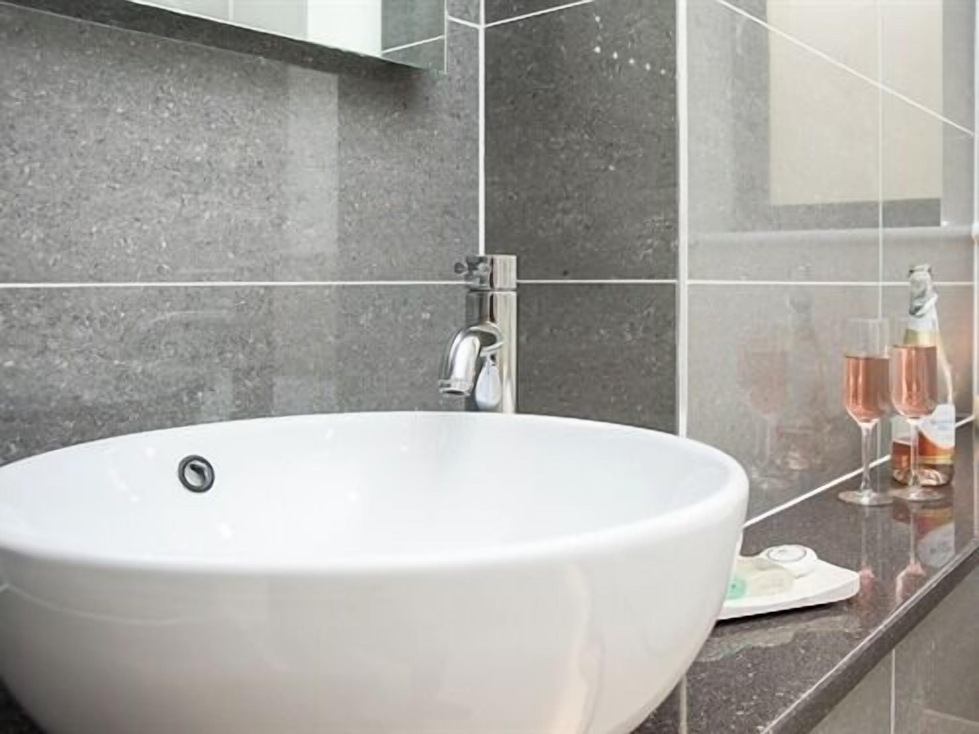 A bathroom with a white sink, a modern faucet, and a mirror above it. There are two glasses and a bottle on the countertop.
