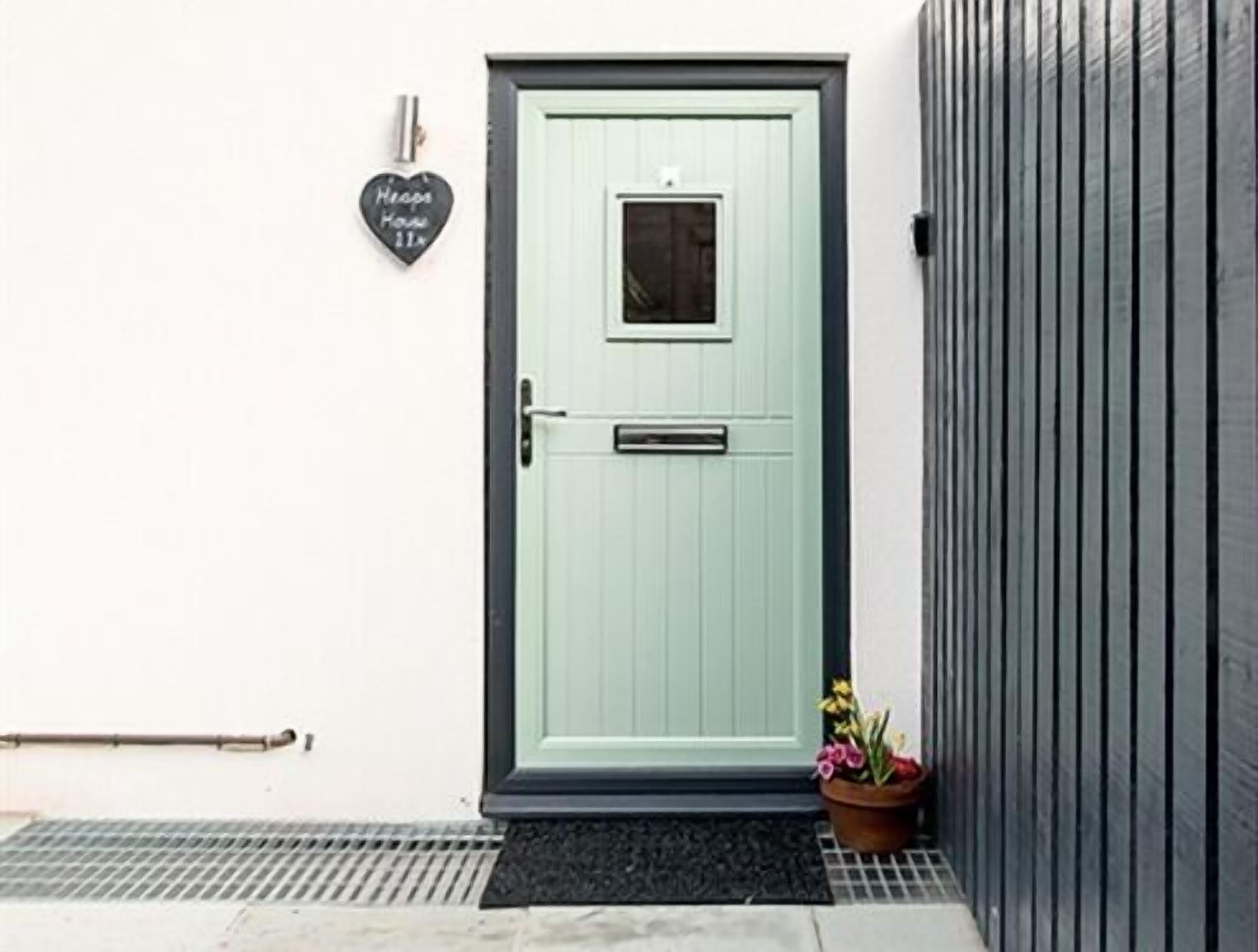 A light green door with a small window, a heart-shaped sign on the wall, a metal handrail, and a potted plant with flowers.