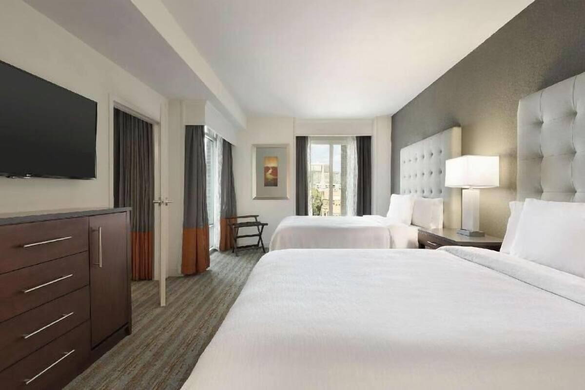 Ein Hotelzimmer mit zwei Betten, einem Flachbildfernseher, einem Schreibtisch, einem Sessel und einem Fenster mit Vorhängen.