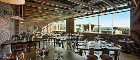 En rymlig restaurang med högt i tak, synliga takbjälkar och stora fönster som erbjuder utsikt över stadsbilden.